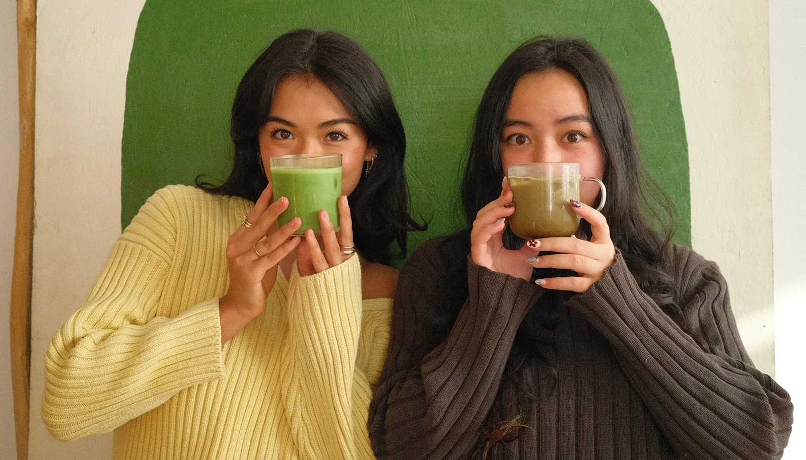 Two people standing against a green painted wall, each holding a glass of matcha up in front of their face