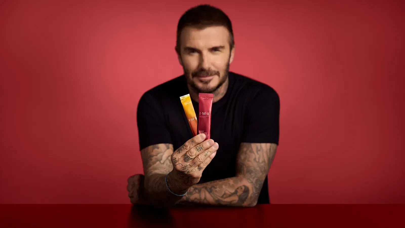 David Beckham in a black t-shirt holding two IM8 sachets against a red background