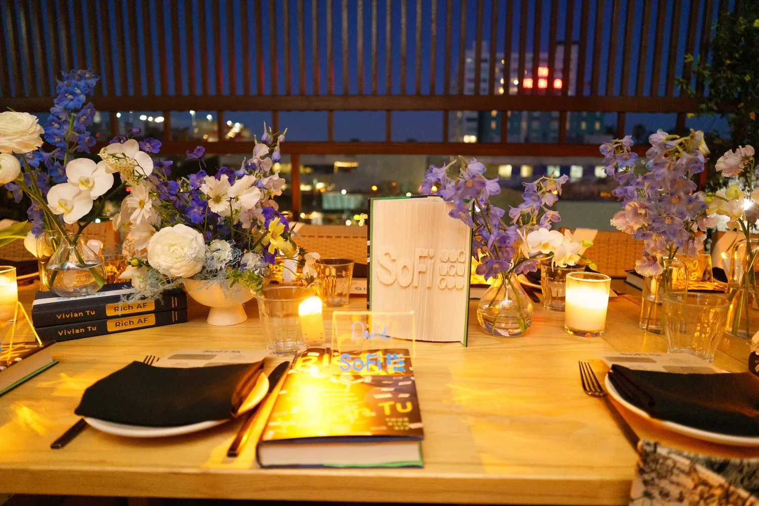The SoFi dinner table at Nasdaq, set with candles, flowers and stacked copies of Vivian Tu's book Rich AF, with the New York skyline behind it at dusk