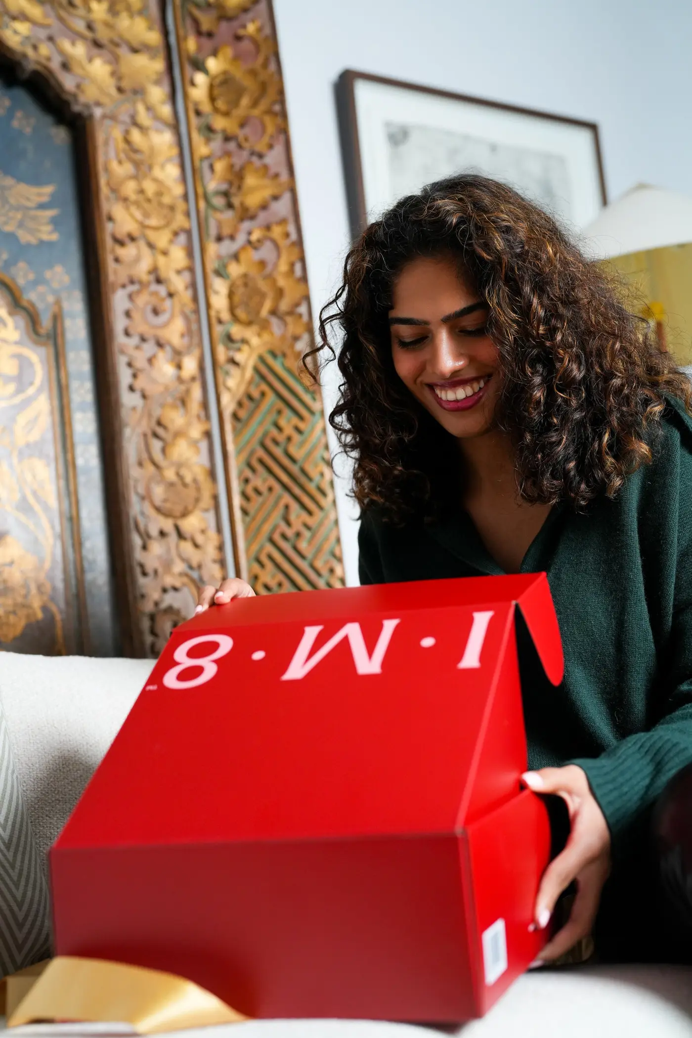 A woman smiling as she opens a large red 'I.M.8' branded gift box on a couch