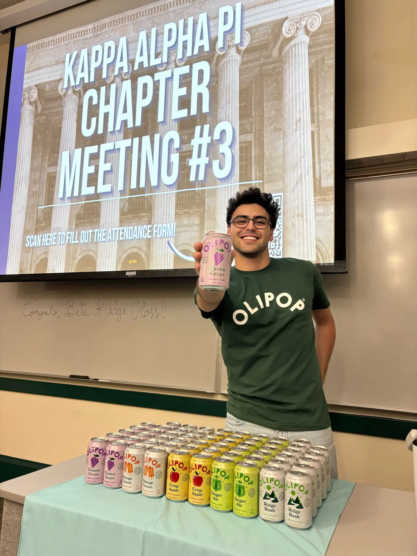 An ambassador in an OLIPOP tee holding a can, presenting in front of a slide reading 'Kappa Alpha Pi Chapter Meeting #3', with roughly 60 cans across 5+ flavors laid out on the table