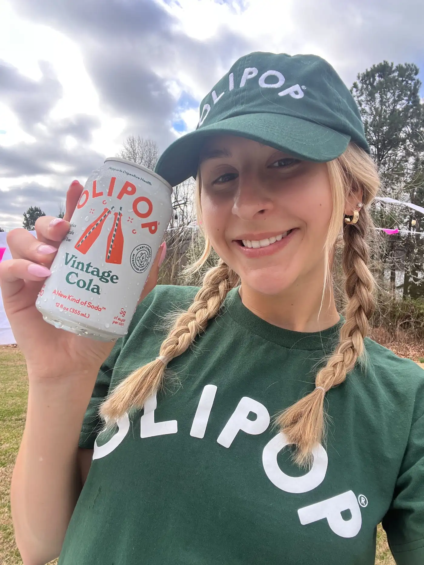 An ambassador outdoors wearing a full OLIPOP branded cap-and-tee kit, smiling and holding a can of Vintage Cola