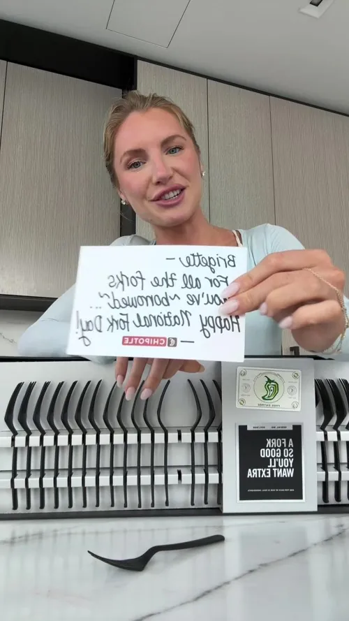 Brigette Pheloung holding the handwritten note over a long white case of black Chipotle forks, each in its own slot, with a free entree card in the middle. Filmed on a front camera, so the writing reads backwards