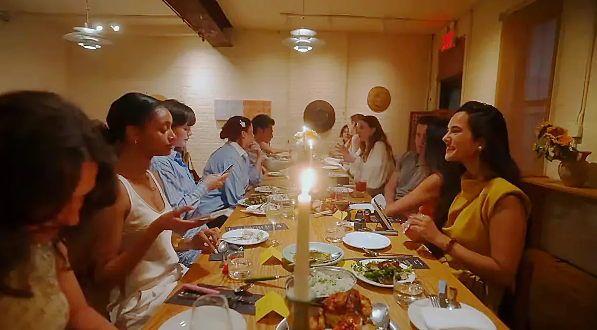 A long table of creators eating, talking and filming by candlelight